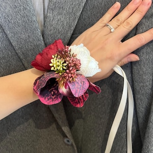 May include: A floral wrist corsage featuring a large dark red peony, a white peony, and a spotted purple and black orchid. The arrangement includes small clusters of green and reddish-brown filler flowers, secured with a long white satin ribbon.