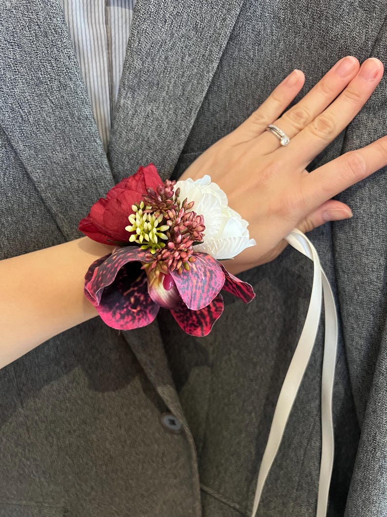 May include: A floral wrist corsage featuring a large dark red peony, a white peony, and a spotted purple and black orchid. The arrangement includes small clusters of green and reddish-brown filler flowers, secured with a long white satin ribbon.