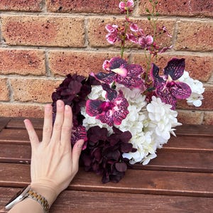 May include: A floral arrangement featuring deep purple and white hydrangea blossoms mixed with vibrant purple and pink speckled orchids. The bouquet displays a dense cluster of petals with tall, slender stems extending upward from the center.