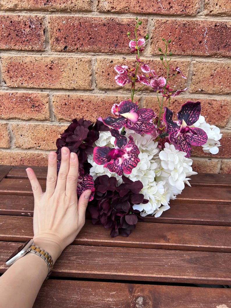 May include: A floral arrangement featuring deep purple and white hydrangea blossoms mixed with vibrant purple and pink speckled orchids. The bouquet displays a dense cluster of petals with tall, slender stems extending upward from the center.
