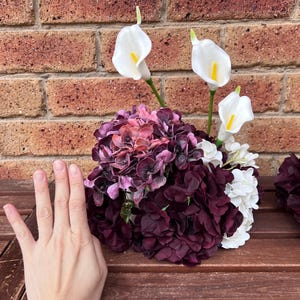 May include: A floral arrangement featuring deep purple and dark pink hydrangea blossoms mixed with white hydrangea clusters and three white calla lilies with yellow centers. The arrangement is displayed as a rounded bouquet of artificial flowers.