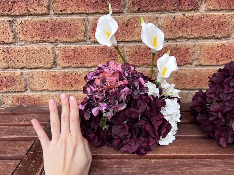 May include: A floral arrangement featuring deep purple and dark pink hydrangea blossoms mixed with white hydrangea clusters and three white calla lilies with yellow centers. The arrangement is displayed as a rounded bouquet of artificial flowers.