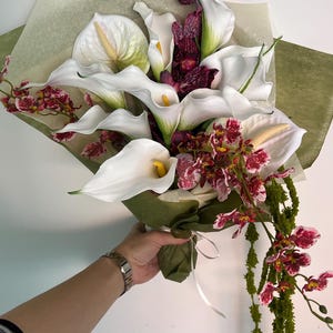May include: A floral bouquet featuring white calla lilies with yellow spadices, dark purple and white mottled orchids, and long, trailing green amaranth stems. The flowers are wrapped in translucent light green paper and tied with a white ribbon.