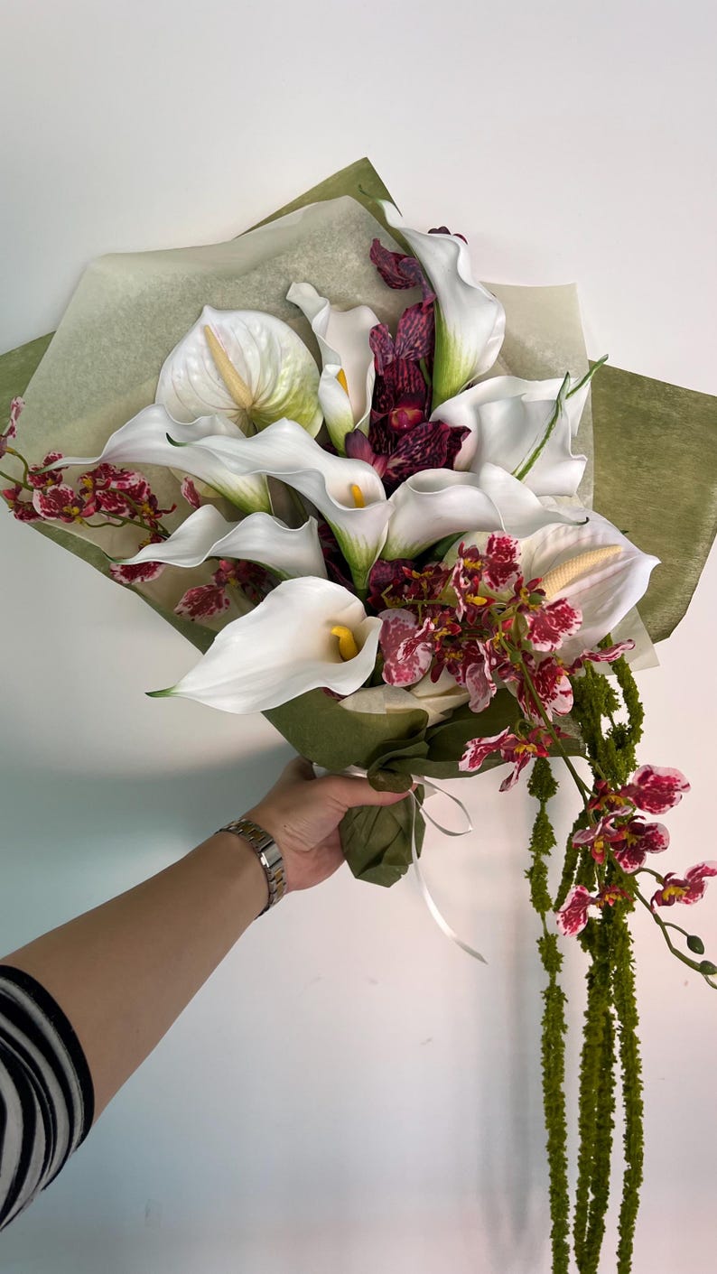 May include: A floral bouquet featuring white calla lilies with yellow spadices, dark purple and white mottled orchids, and long, trailing green amaranth stems. The flowers are wrapped in translucent light green paper and tied with a white ribbon.