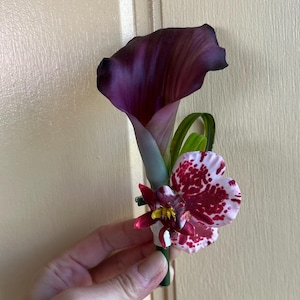 May include: A floral boutonniere featuring a deep purple calla lily paired with a white orchid petal displaying dark red speckles and a smaller dark red flower accent with a yellow center, all secured on a green stem base.