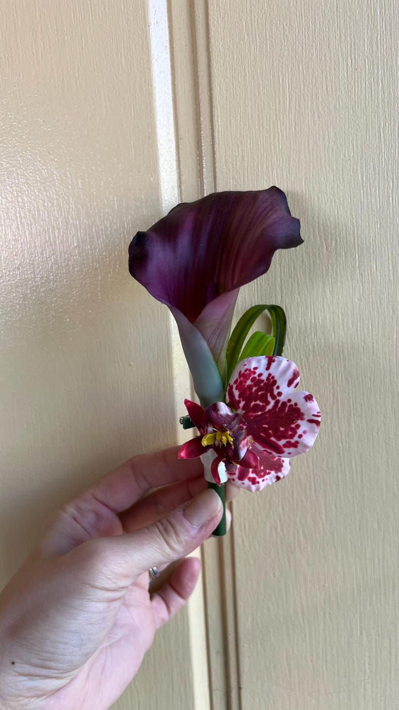 May include: A floral boutonniere featuring a deep purple calla lily paired with a white orchid petal displaying dark red speckles and a smaller dark red flower accent with a yellow center, all secured on a green stem base.