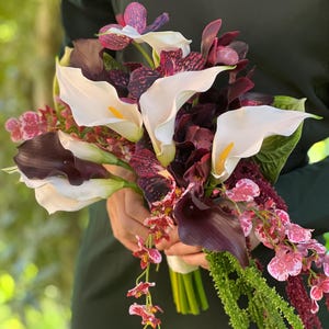 May include: A floral bouquet featuring white calla lilies with yellow centers, deep purple calla lilies, dark purple patterned orchids, and small pink and white speckled flowers, accented with long green hanging amaranth stems and green leaves.