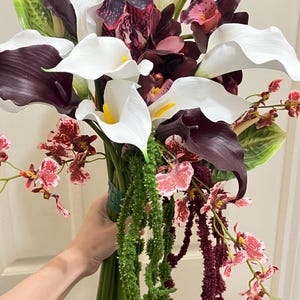 May include: A floral bouquet featuring white and deep purple calla lilies, dark burgundy orchids with speckled patterns, and small pink and red patterned orchids. The arrangement includes long, trailing green and dark red amaranth stems.