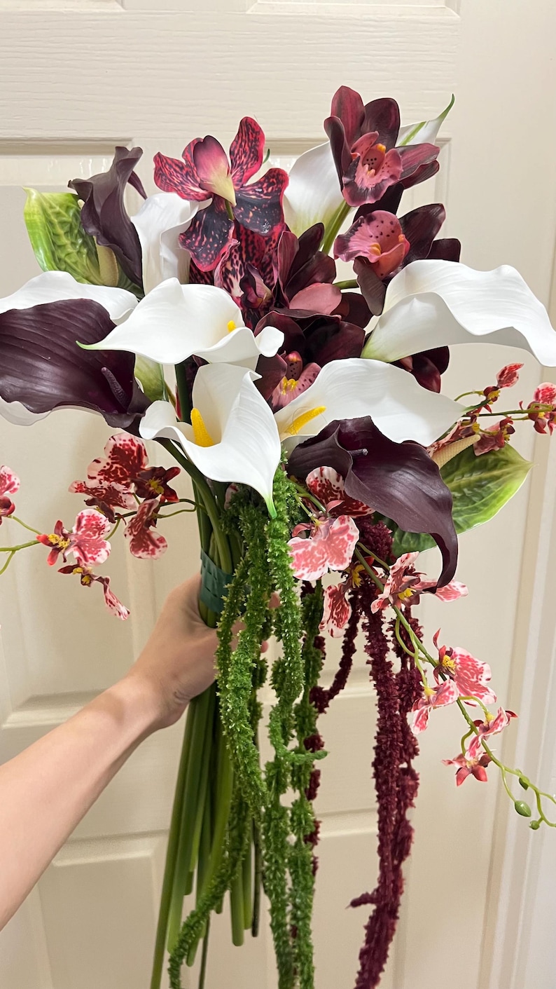 May include: A floral bouquet featuring white and deep purple calla lilies, dark burgundy orchids with speckled patterns, and small pink and red patterned orchids. The arrangement includes long, trailing green and dark red amaranth stems.