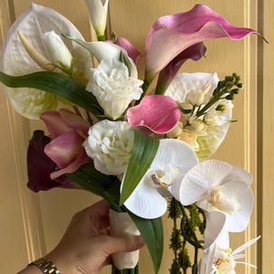 May include: A bouquet of artificial flowers featuring white calla lilies, pink and purple calla lilies, white orchids, and white roses. The bouquet is wrapped with white ribbon and green leaves.