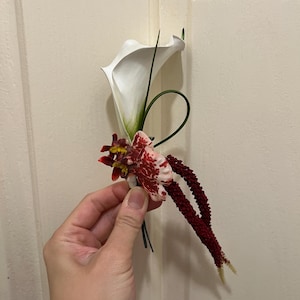 May include: A floral boutonniere featuring a single white calla lily, a small mottled red and white orchid, and two long, textured dark burgundy amaranth stems. The arrangement includes a thin, curved green grass blade and dark wire stems.