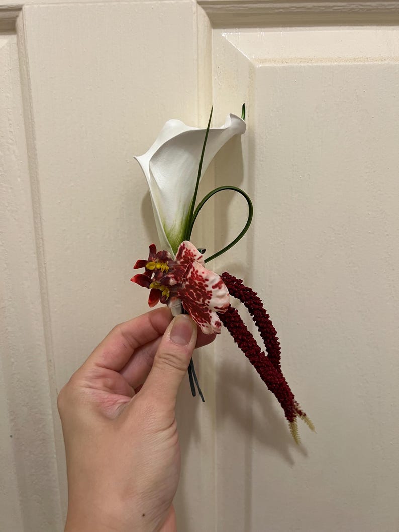 May include: A floral boutonniere featuring a single white calla lily, a small mottled red and white orchid, and two long, textured dark burgundy amaranth stems. The arrangement includes a thin, curved green grass blade and dark wire stems.