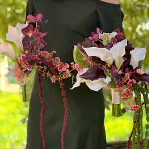 May include: Two floral bouquets featuring white calla lilies, deep purple calla lilies, dark maroon orchids with speckled patterns, and long, trailing green and maroon amaranth stems. Each bouquet is bound with a white ribbon at the handle.