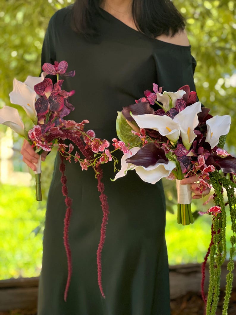 May include: Two floral bouquets featuring white calla lilies, deep purple calla lilies, dark maroon orchids with speckled patterns, and long, trailing green and maroon amaranth stems. Each bouquet is bound with a white ribbon at the handle.