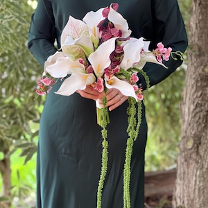May include: A floral bouquet featuring white calla lilies with yellow spadices, deep burgundy mottled orchids, and long, trailing green amaranth stems. The arrangement is held together by a white ribbon wrap, creating a cascading botanical display.