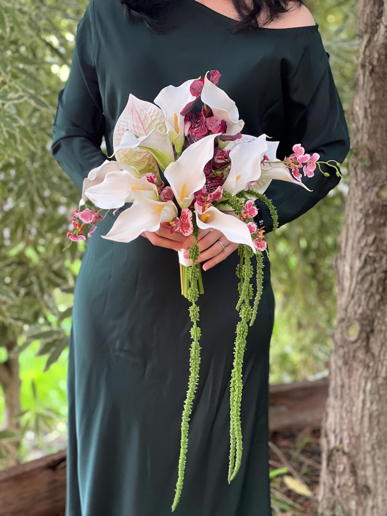May include: A floral bouquet featuring white calla lilies with yellow spadices, deep burgundy mottled orchids, and long, trailing green amaranth stems. The arrangement is held together by a white ribbon wrap, creating a cascading botanical display.