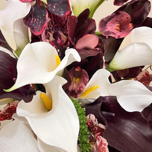 May include: A floral arrangement featuring white calla lilies with yellow spadices, deep burgundy orchids with speckled patterns, and white phalaenopsis orchids. The bouquet includes a mix of dark purple and white petals with green accents.