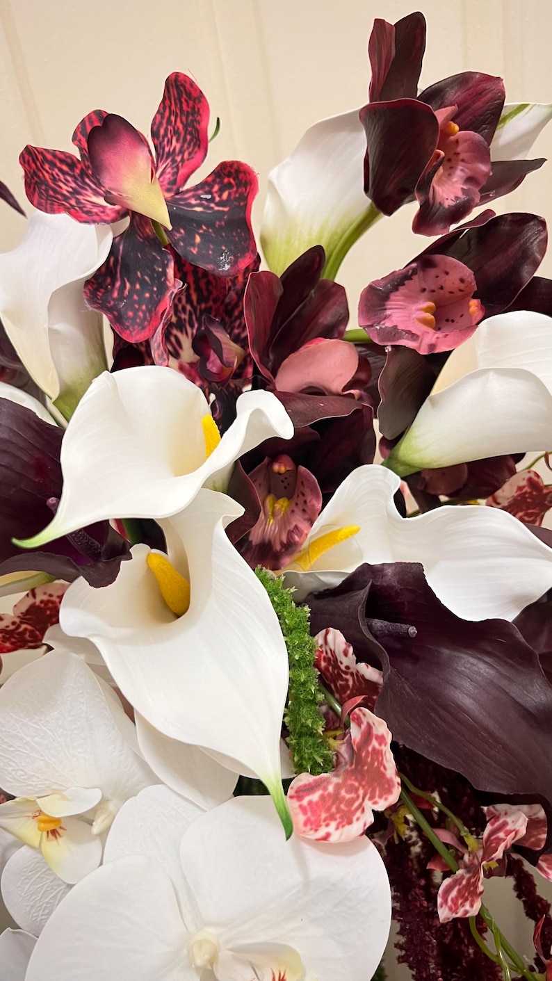May include: A floral arrangement featuring white calla lilies with yellow spadices, deep burgundy orchids with speckled patterns, and white phalaenopsis orchids. The bouquet includes a mix of dark purple and white petals with green accents.
