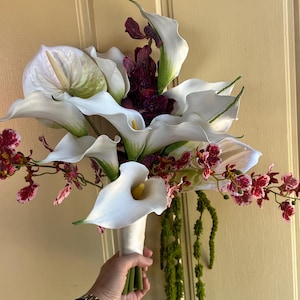 May include: A floral bouquet featuring large white calla lilies with yellow spadices, deep purple speckled orchids, and long trailing green amaranthus. The stems are bound together with a smooth white ribbon wrap at the base of the arrangement.