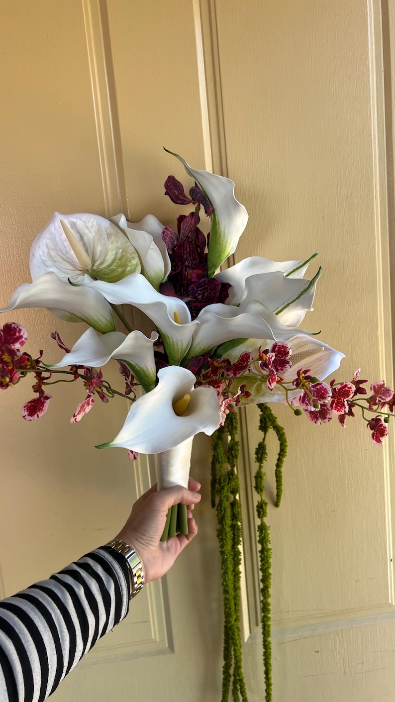 May include: A floral bouquet featuring large white calla lilies with yellow spadices, deep purple speckled orchids, and long trailing green amaranthus. The stems are bound together with a smooth white ribbon wrap at the base of the arrangement.
