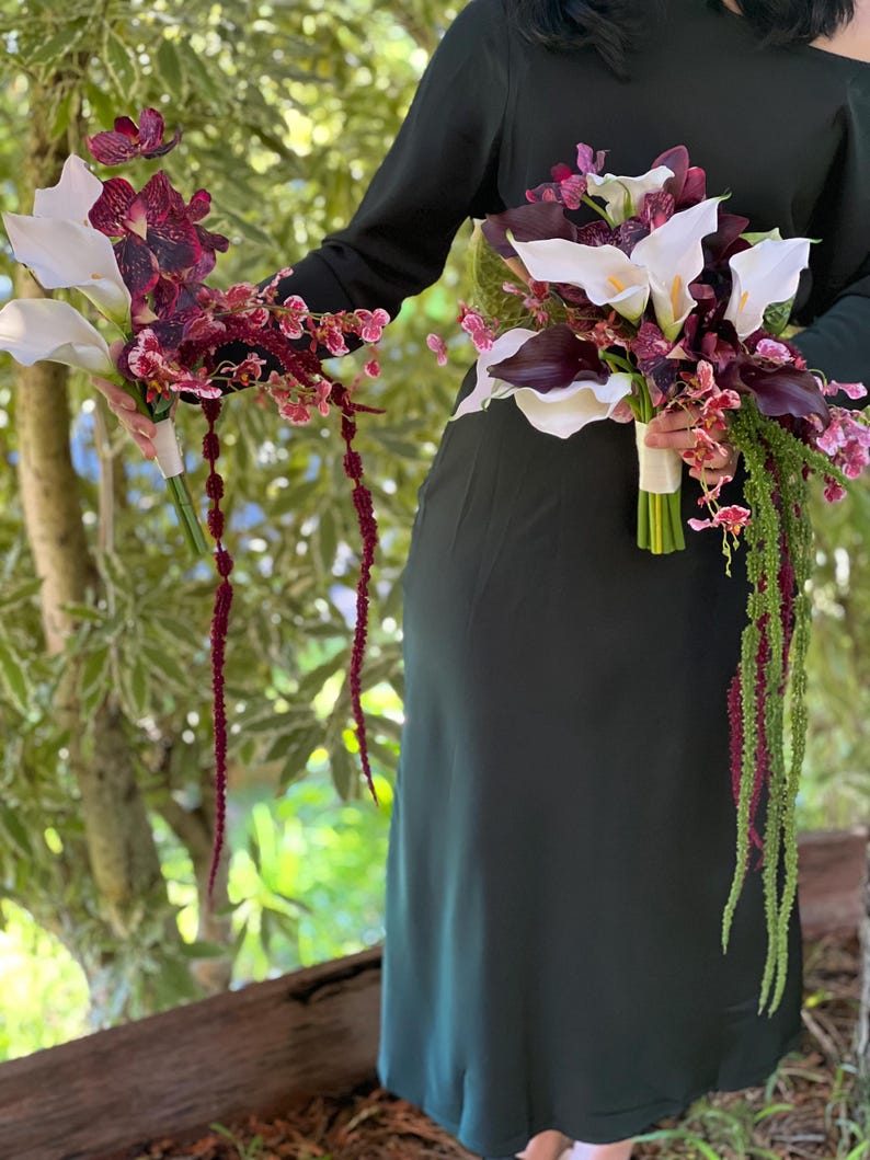 May include: Two floral bouquets featuring white calla lilies, deep purple orchids, and cascading amaranth stems. The bouquets are wrapped in white ribbon at the stems. The arrangement includes a mix of dark burgundy and white floral elements.