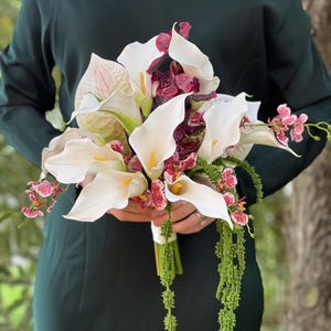 May include: A floral bouquet featuring white calla lilies with yellow centers, deep burgundy patterned orchids, and long, trailing green amaranthus. The arrangement is held together with a white ribbon wrap, creating a structured, multi-textured display.