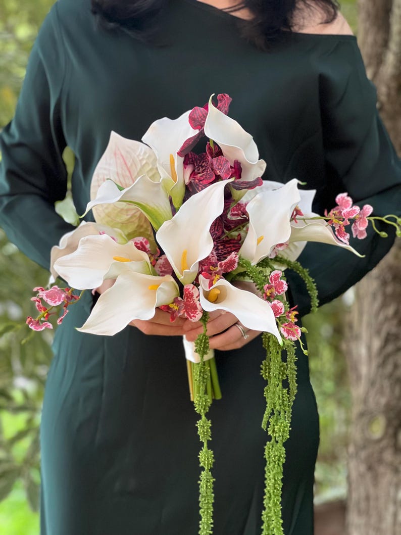 May include: A floral bouquet featuring white calla lilies with yellow centers, deep burgundy patterned orchids, and long, trailing green amaranthus. The arrangement is held together with a white ribbon wrap, creating a structured, multi-textured display.