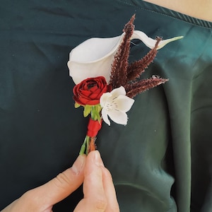 Pu&ograve; includere: Un'occhiello floreale con una calla bianca, una rosa rossa e un fiore bianco con accenti marroni. I fiori sono legati con un nastro rosso. L'occhiello &egrave; tenuto contro un tessuto verde scuro.