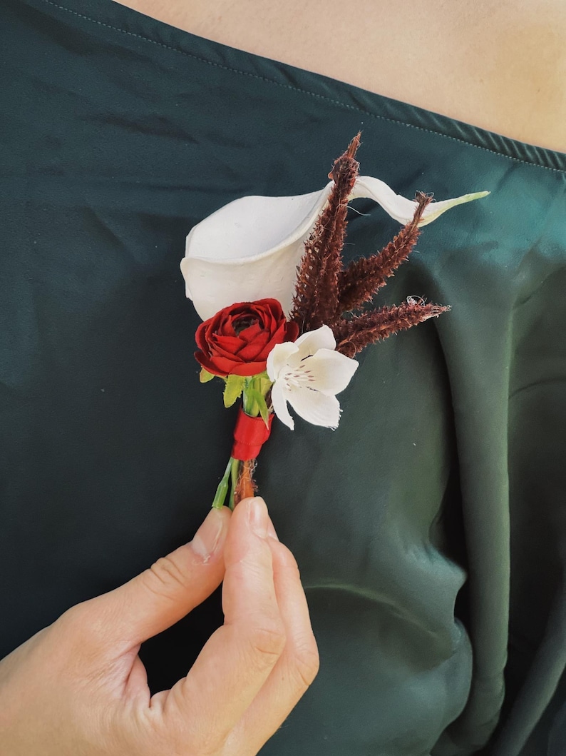 Pu&ograve; includere: Un'occhiello floreale con una calla bianca, una rosa rossa e un fiore bianco con accenti marroni. I fiori sono legati con un nastro rosso. L'occhiello &egrave; tenuto contro un tessuto verde scuro.