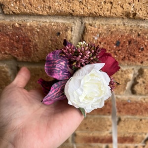 May include: A floral corsage featuring a large white peony, a deep purple orchid with dark speckled patterns, and small clusters of burgundy buds. The arrangement includes thin, light-colored ribbon streamers hanging from the base of the flowers.