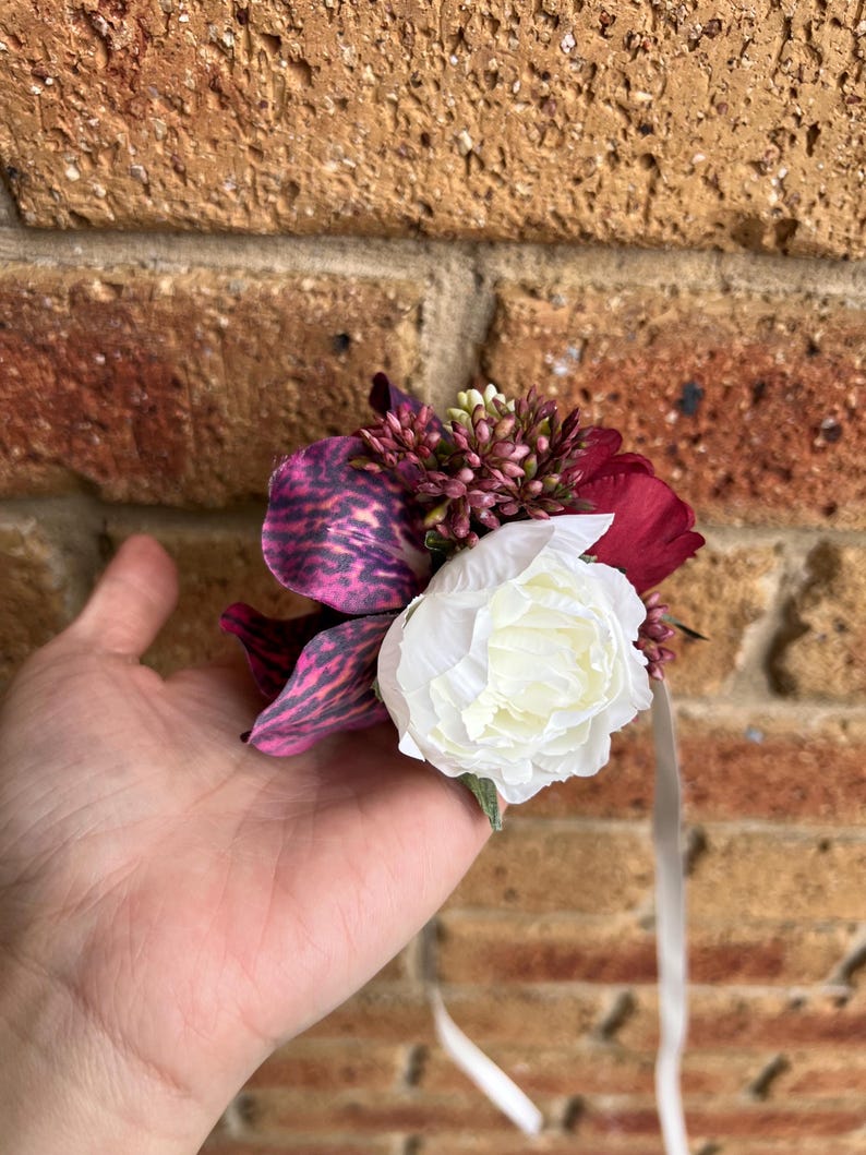 May include: A floral corsage featuring a large white peony, a deep purple orchid with dark speckled patterns, and small clusters of burgundy buds. The arrangement includes thin, light-colored ribbon streamers hanging from the base of the flowers.