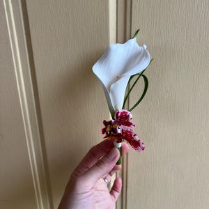 May include: A boutonniere featuring a single white calla lily flower paired with a smaller orchid blossom that has deep red and white mottled petals. The floral arrangement is accented with a thin green stem and secured with white floral tape.