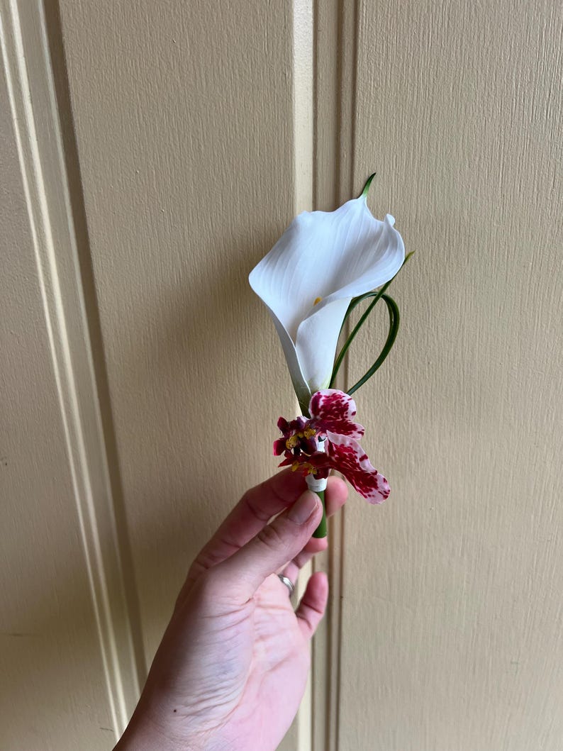 May include: A boutonniere featuring a single white calla lily flower paired with a smaller orchid blossom that has deep red and white mottled petals. The floral arrangement is accented with a thin green stem and secured with white floral tape.