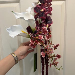 May include: A floral bouquet featuring two large white calla lilies with yellow centers, several deep burgundy and purple mottled orchids, smaller white and red speckled orchids, and long, thin, dark red hanging amaranth stems.