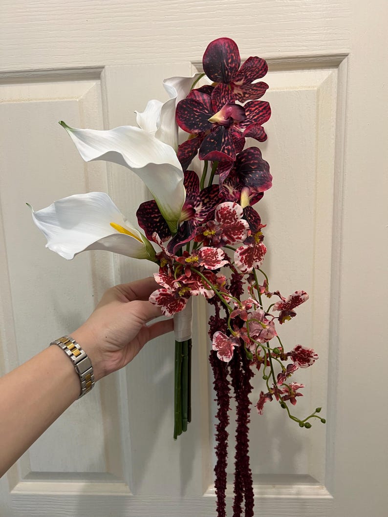 May include: A floral bouquet featuring two large white calla lilies with yellow centers, several deep burgundy and purple mottled orchids, smaller white and red speckled orchids, and long, thin, dark red hanging amaranth stems.