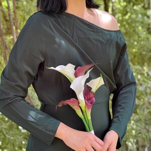 Pu&ograve; includere: Un abito verde scuro a maniche lunghe con scollatura asimmetrica. &Egrave; abbinato a un bouquet di calle bianche e bordeaux, legate con un nastro bianco. Lo sfondo presenta un fogliame verde sfocato, creando un ambiente naturale.