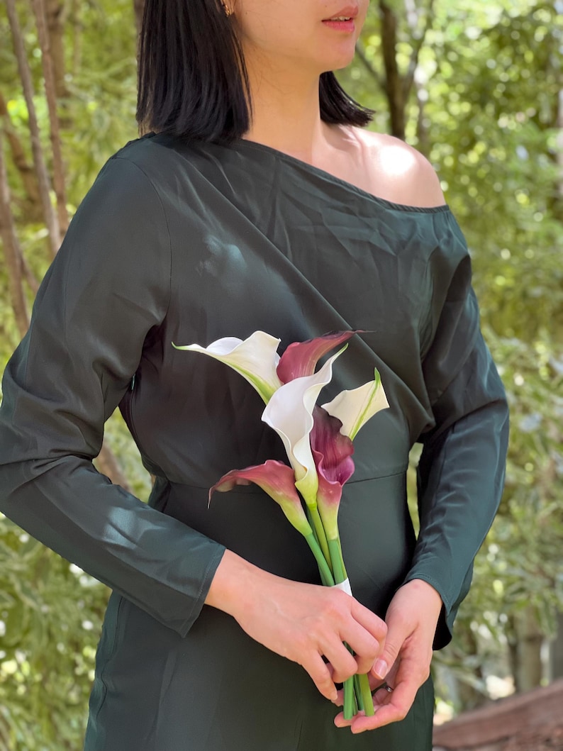 Pu&ograve; includere: Un abito verde scuro a maniche lunghe con scollatura asimmetrica. &Egrave; abbinato a un bouquet di calle bianche e bordeaux, legate con un nastro bianco. Lo sfondo presenta un fogliame verde sfocato, creando un ambiente naturale.