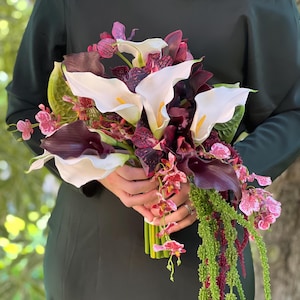 May include: A floral bouquet featuring white calla lilies, deep purple calla lilies, and dark purple speckled orchids. The arrangement includes cascading green amaranth and small pink and white patterned orchid blossoms, tied with a green stem wrap.