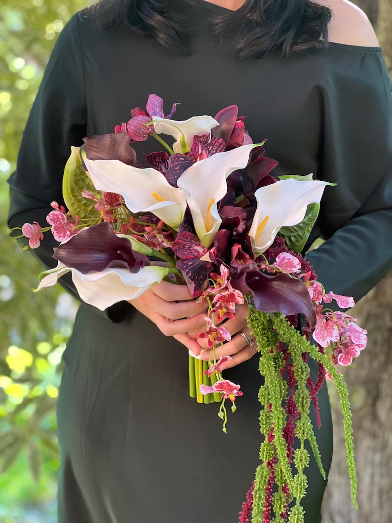 May include: A floral bouquet featuring white calla lilies, deep purple calla lilies, and dark purple speckled orchids. The arrangement includes cascading green amaranth and small pink and white patterned orchid blossoms, tied with a green stem wrap.