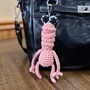 May include: A handmade pink crochet keychain in the shape of a whimsical creature with black and white eyes. The keychain is attached to a black leather handbag with silver hardware. The background is a wooden surface.