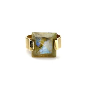 May include: A gold ring with a square-shaped labradorite gemstone. The gemstone has a light green, blue, and gray color.