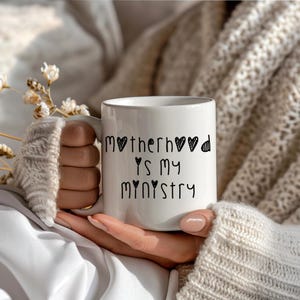 May include: White ceramic mug with the text "Motherhood is my Ministry" in black script. The mug is held by a person wearing a cream-colored sweater. The mug has a standard handle and a glossy finish.