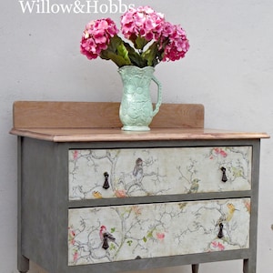 May include: A gray painted two-drawer dresser with a wood top. The drawers have a floral print with birds. The dresser is sitting on a wood floor.  Willow & Hobbs is written on the wall behind the dresser.