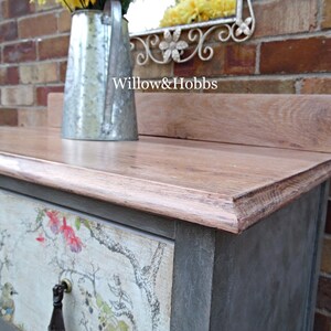 May include: A gray painted dresser with a wood top and a floral patterned drawer. The dresser has a metal handle and is sitting in front of a brick wall. The text "Willow & Hobbs" is visible on the top of the dresser.