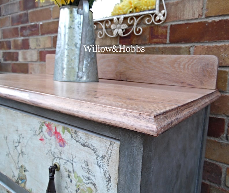 May include: A gray painted dresser with a wood top and a floral patterned drawer. The dresser has a metal handle and is sitting in front of a brick wall. The text "Willow & Hobbs" is visible on the top of the dresser.