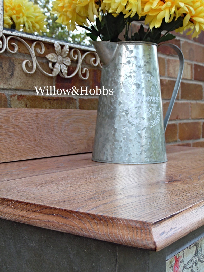 May include: A rustic wooden table top with a distressed gray finish. A galvanized metal pitcher with a floral design and the word "Lavender" is sitting on the table. The pitcher is filled with yellow flowers.