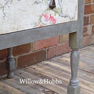May include: A gray painted wooden side table with a floral design on the top drawer. The table has four turned legs and a gray painted base.
