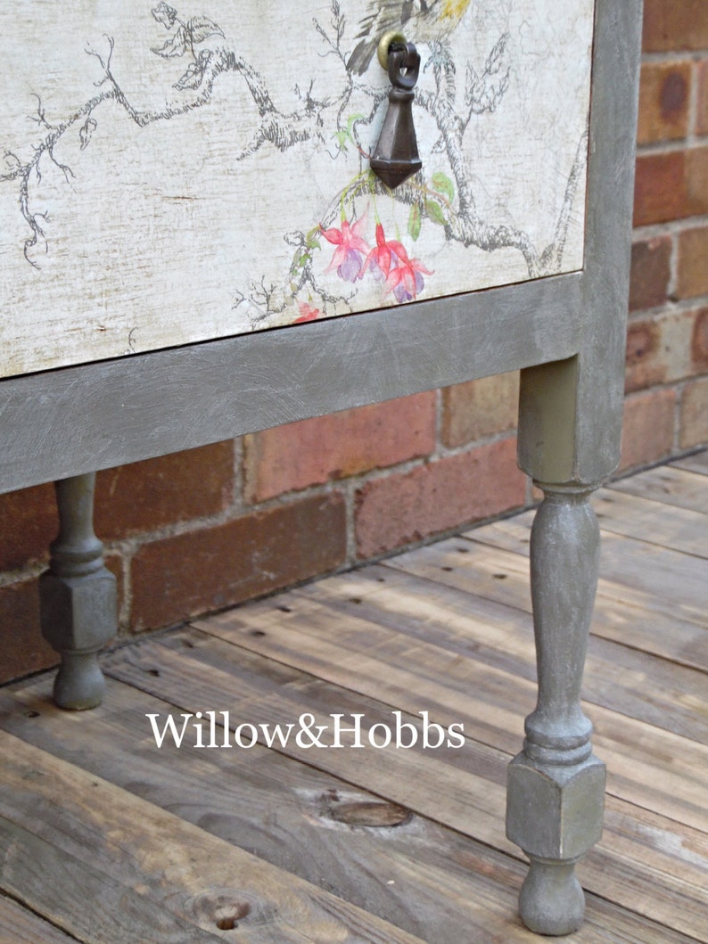 May include: A gray painted wooden side table with a floral design on the top drawer. The table has four turned legs and a gray painted base.