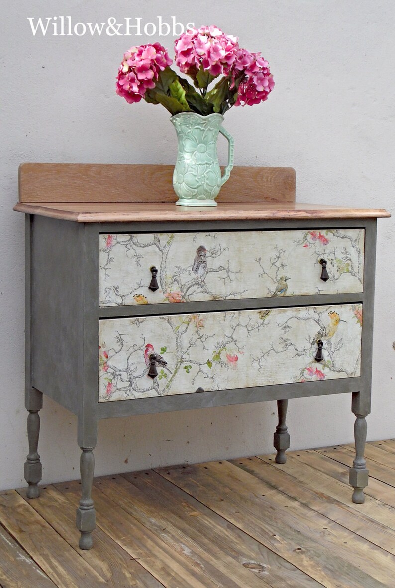 May include: A gray painted two-drawer dresser with a wood top. The drawers have a floral print with birds. The dresser is sitting on a wood floor.  Willow & Hobbs is written on the wall behind the dresser.