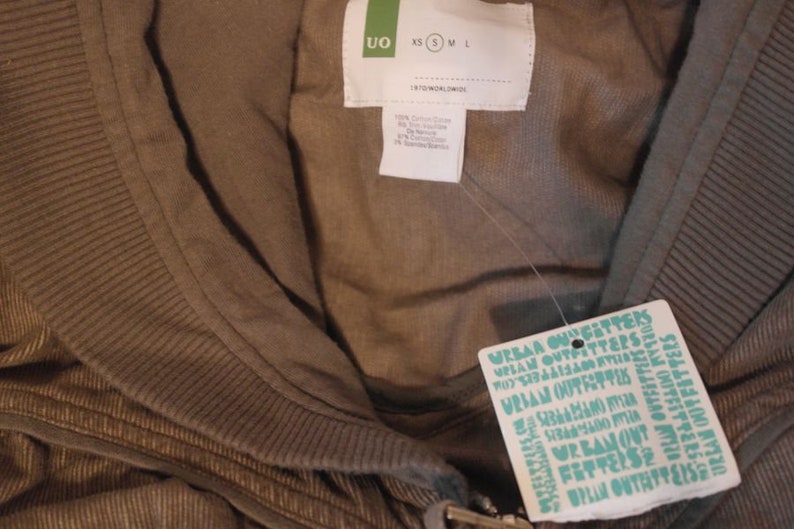 May include: A brown corduroy hoodie with a zipper closure and a tag that reads "Urban Outfitters". The tag is white with teal blue text. The hoodie has a ribbed collar and a size tag that reads "XS S M L".